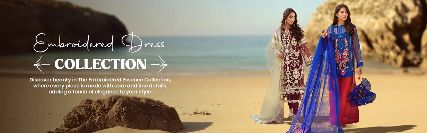 Two women in embroidered dresses stand on a sandy beach, modeling stunning embroidered dress designs from a The Woven Palace, with rocky cliffs and a clear sky in the background.