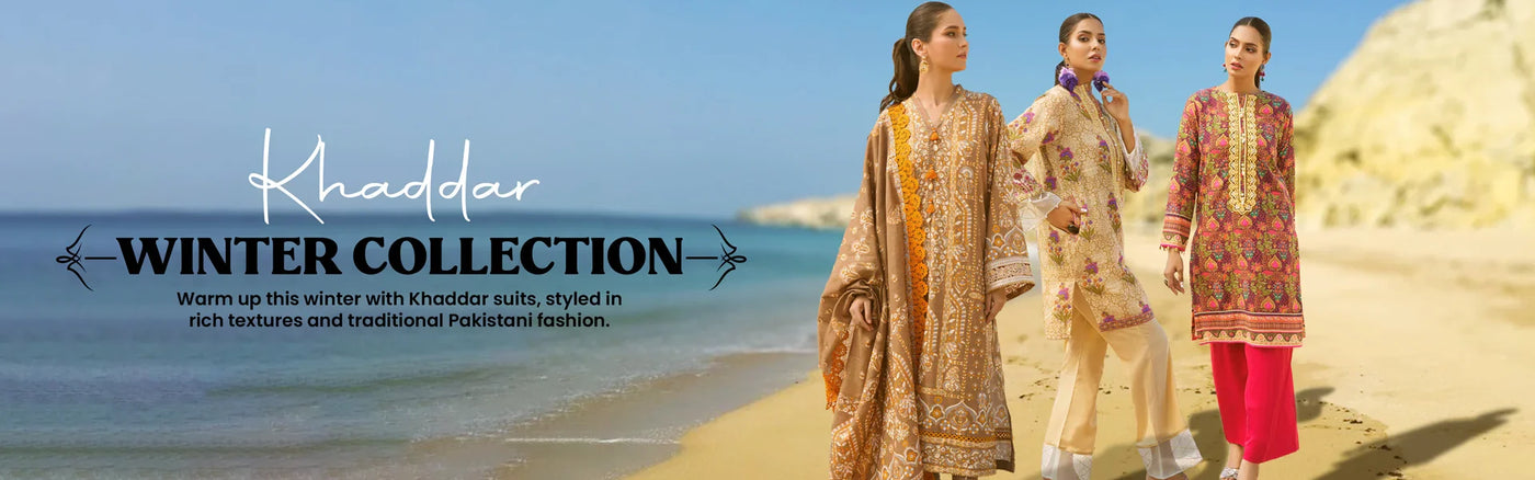 Three women wearing colorful traditional Pakistani khaddar suits walk along a sandy beach with rocky cliffs in the background.